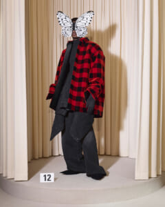 Model on a runway wearing a red and black plaid oversized jacket over black layers, head obscured by a white butterfly mask; stage with beige curtains behind.