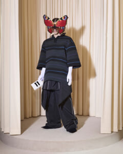 Model on a beige-curtained stage wearing a dark striped oversized top, white gloves, and layered black bottoms, with a large red butterfly mask and a card labeled '11'.