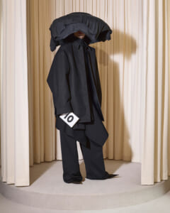 Person in an oversized black suit and large hood stands on a circular platform, holding a white card with the number 10 amid beige stage curtains.