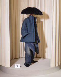 Mannequin in an oversized blue jacket and baggy denim under a large black feather umbrella on a circular display with beige curtains.