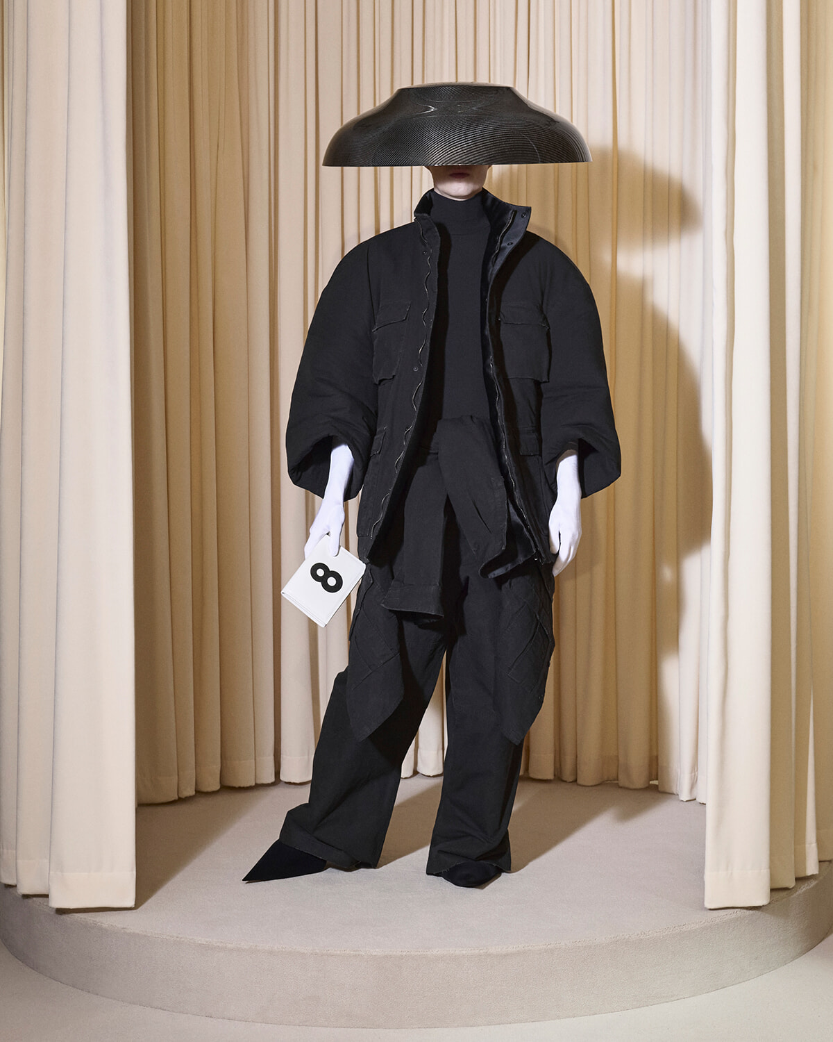 Mannequin in an oversized black layered outfit on a circular pedestal, under a large black lampshade, holding a tag with the number 8.