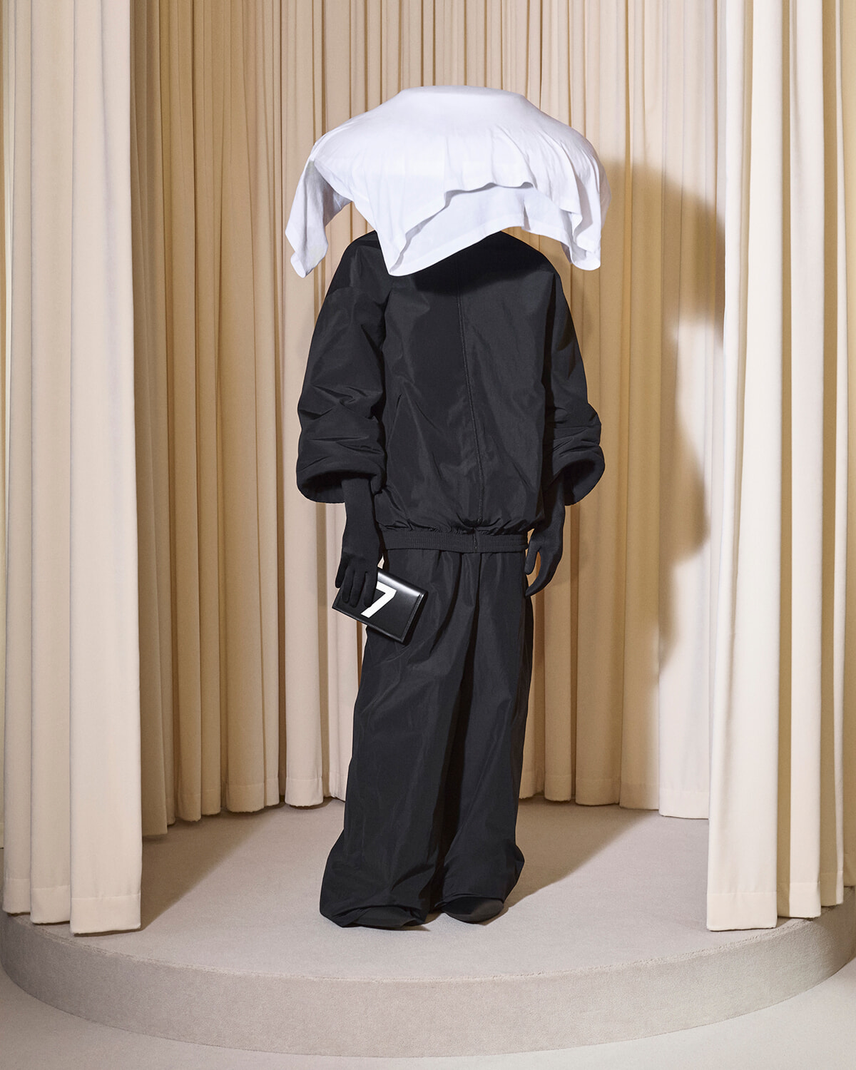 Mannequin wearing a black jumpsuit with a white shirt draped over the head like a hood, on a circular pedestal.