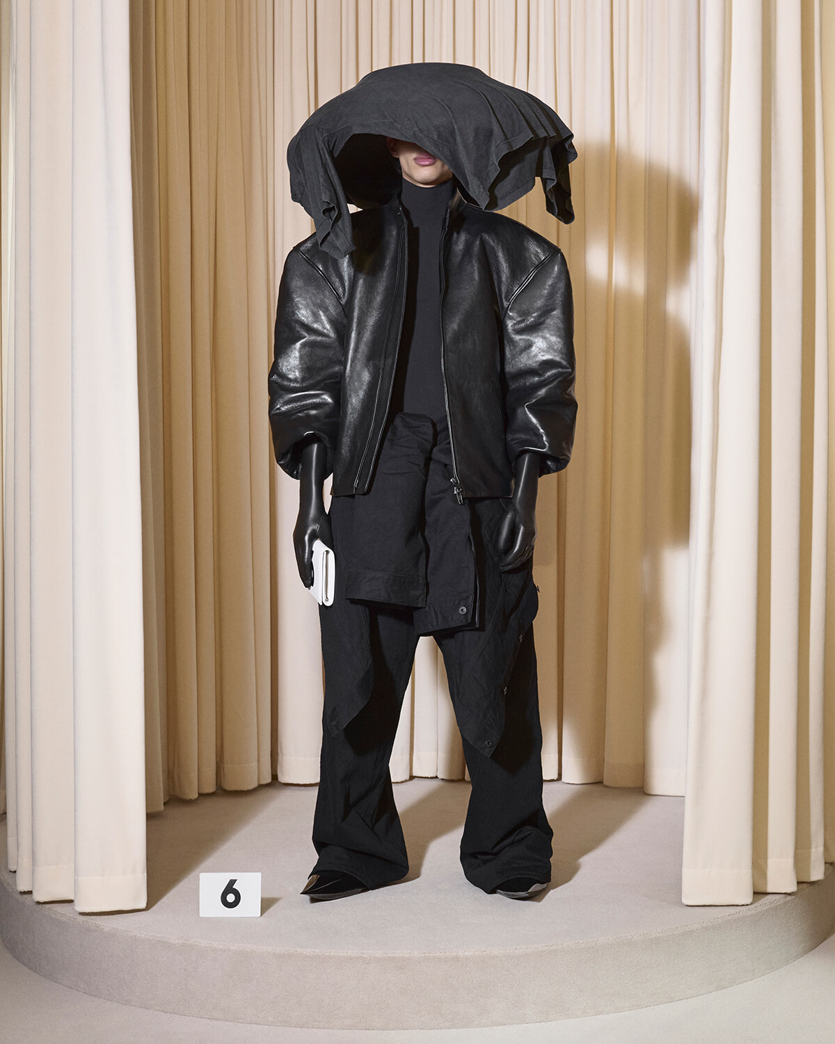 Mannequin in a oversized black leather jacket with a draped hood over the head, standing on a circular runway with curtain backdrop and a white number 6 placard nearby, holding a white clutch.