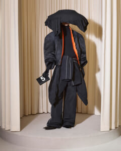 Avant-garde black fashion installation on a circular platform, featuring a large sculptural hat and orange-trim jacket with a small '5' card beside it.
