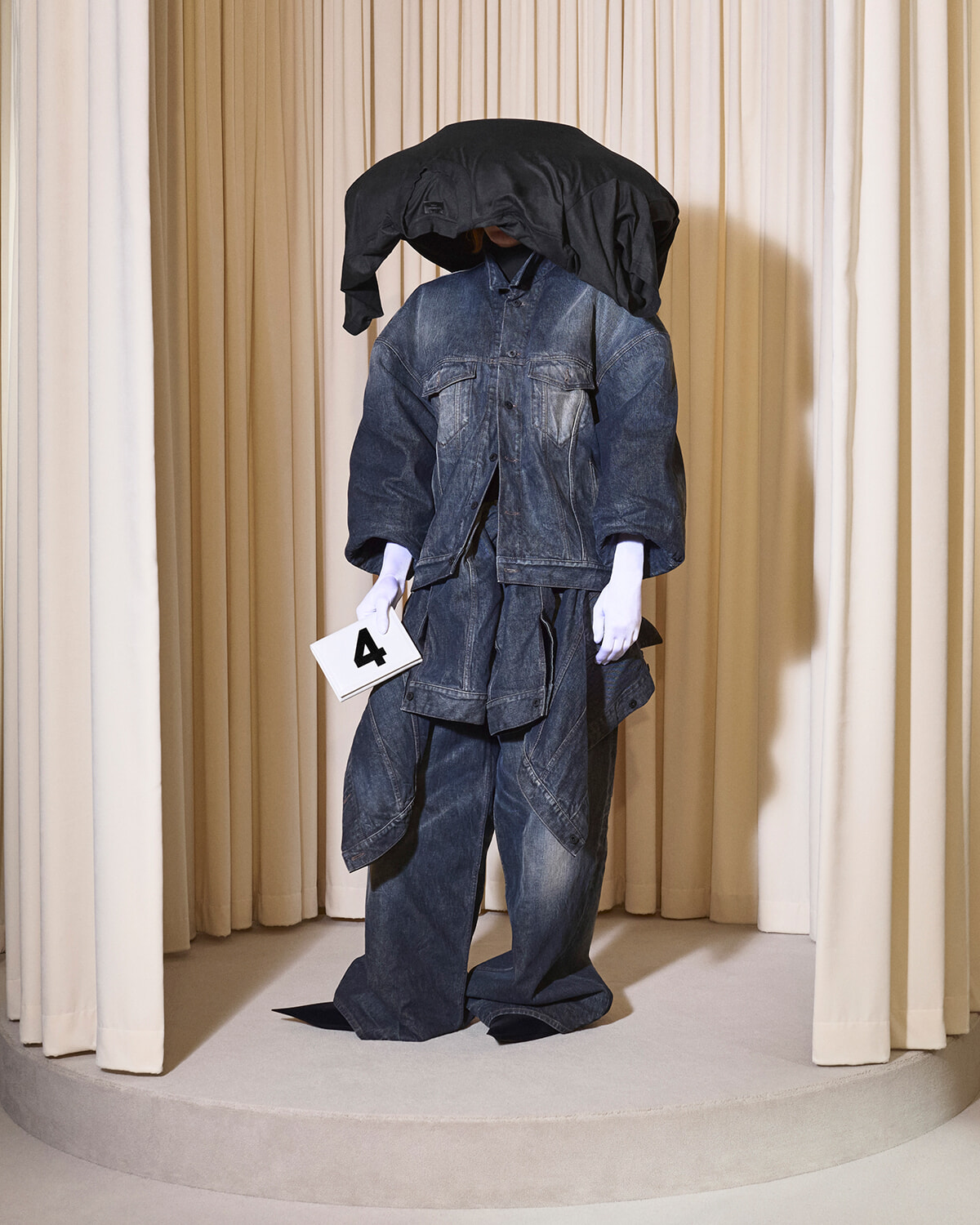Mannequin wearing an oversized denim outfit with a black hood over the head, standing on a circular platform and holding a '4' card.