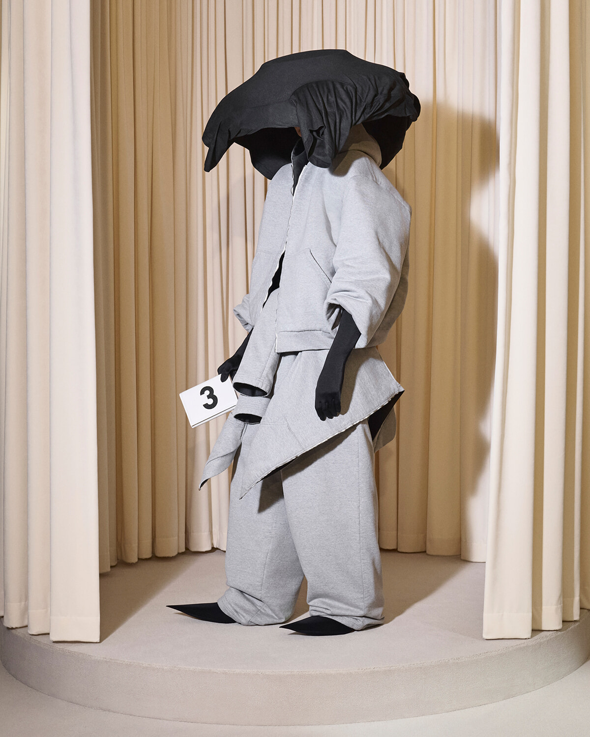 Avant-garde gray layered coat on a mannequin with an oversized black hood, black gloves, standing on a circular beige stage with cream curtains; holding a white number 3 card