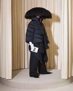 Model on a runway wearing an oversized black puffer with a large fur hood, white gloves, and a numbered card reading '2' in hand.