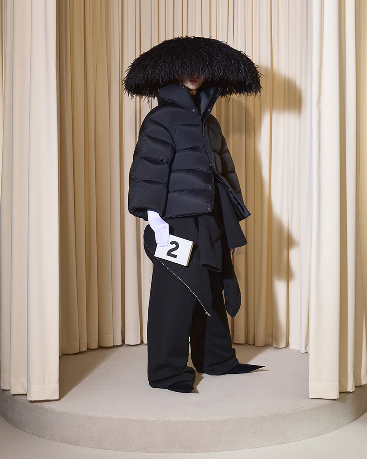 Model on a runway wearing an oversized black puffer with a large fur hood, white gloves, and a numbered card reading '2' in hand.