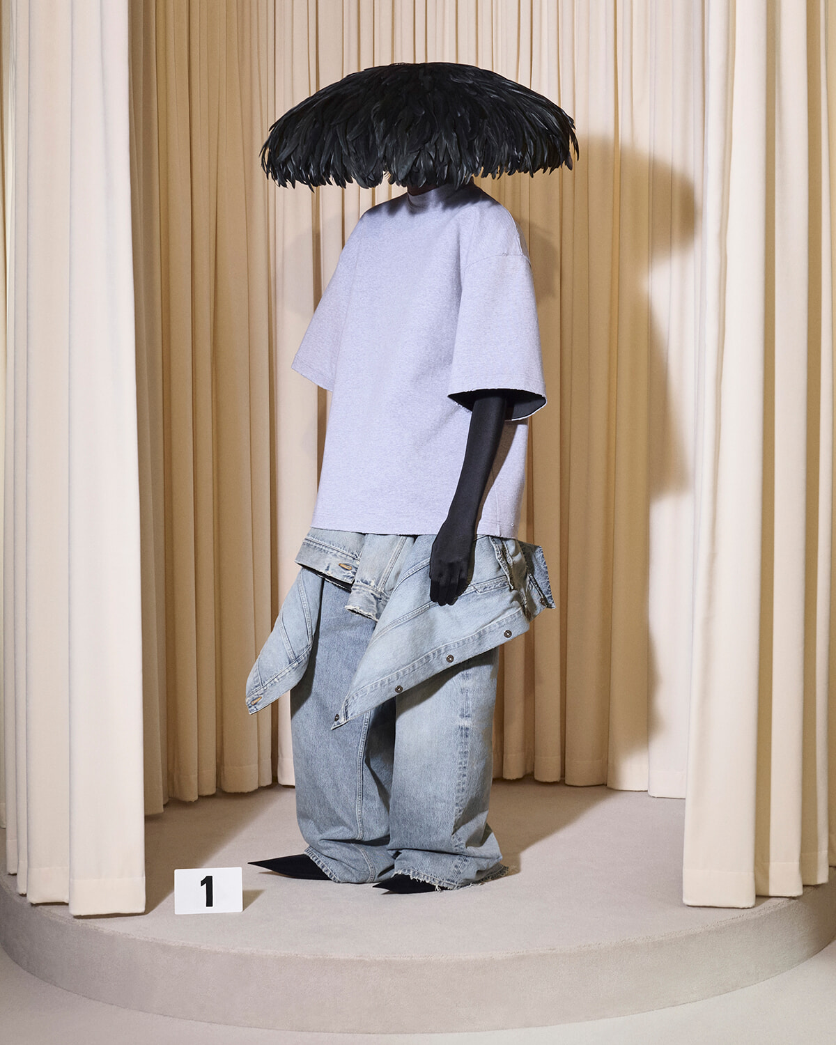Mannequin wearing a lavender T-shirt and oversized jeans, with a denim jacket draped around the hips and a black feathered umbrella hat, on a circular display pedestal marked '1'.