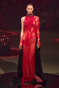 Model on a runway wearing a sheer red lace gown with fringe details and a high-neck beaded collar; a black cape drapes behind her.