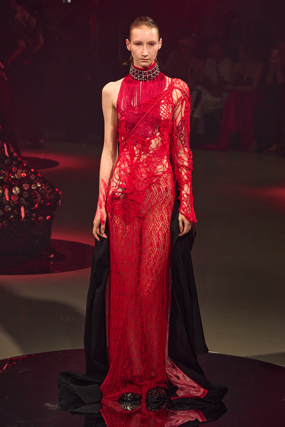 Model on a runway wearing a sheer red lace gown with fringe details and a high-neck beaded collar; a black cape drapes behind her.