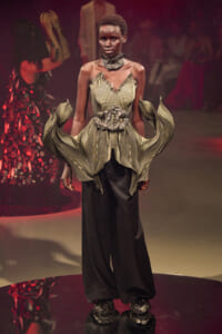 Female model on a runway wearing a sculptural gold-toned metallic gown with leaf-like panels and a jeweled belt.