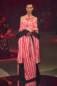 Model on a runway wearing an off-shoulder pink patterned gown with a black sash belt and red paint accents on the face and neck.