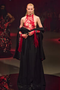 Model on a runway wearing a black floor-length gown with a red crochet bodice and red fringe accents.