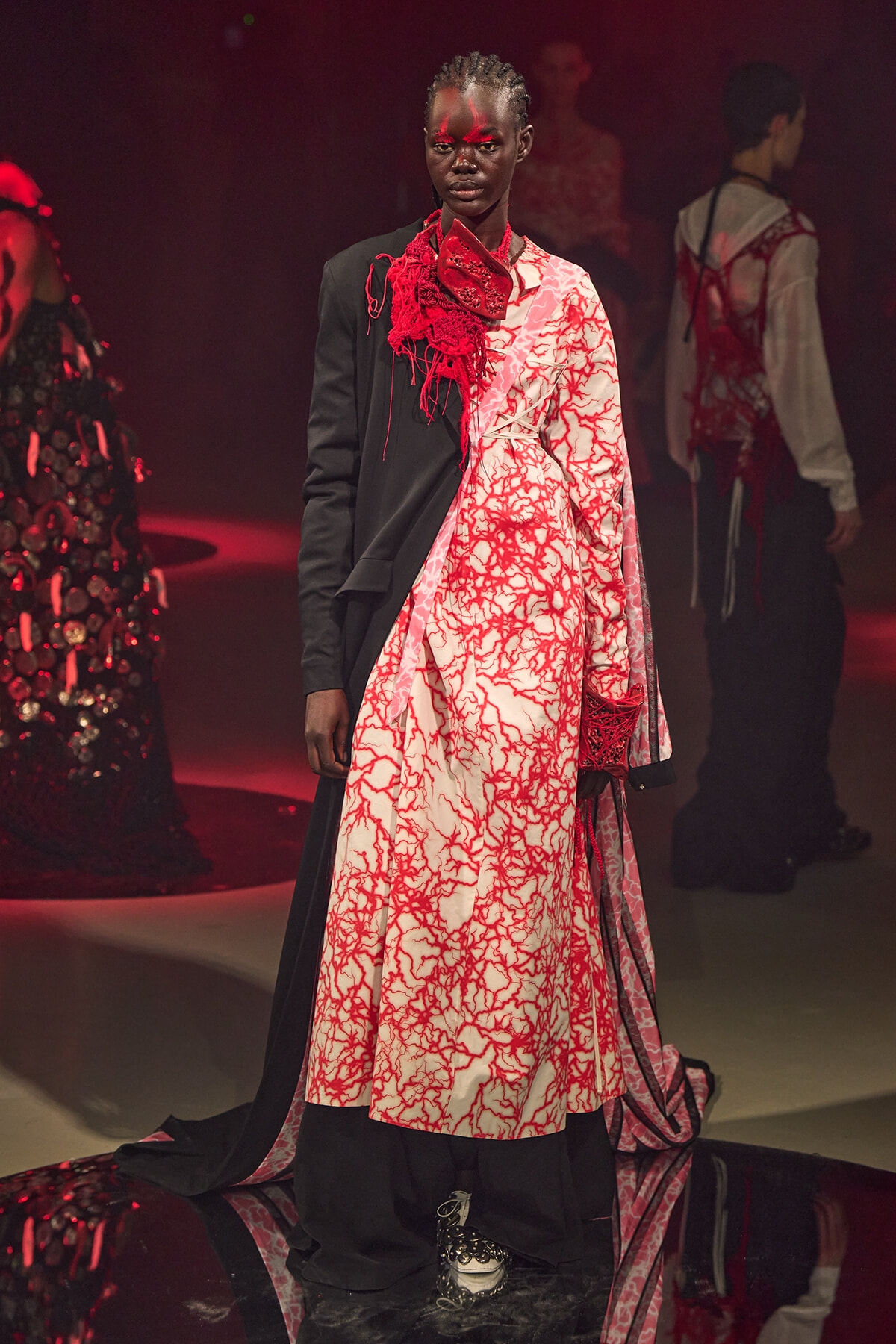 Model on a red-lit runway wearing a split black/white gown with a red vein-like pattern and sculptural neckpiece.