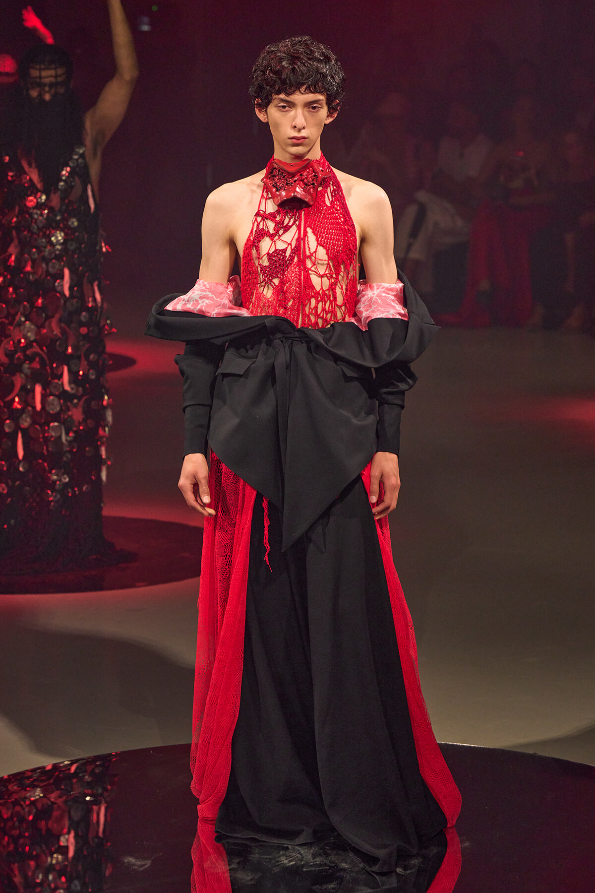 Model walks a fashion runway in a striking red lace halter bodysuit paired with a large black wrapped overlay and pink-printed cuffs, spotlight on a high-fashion look.