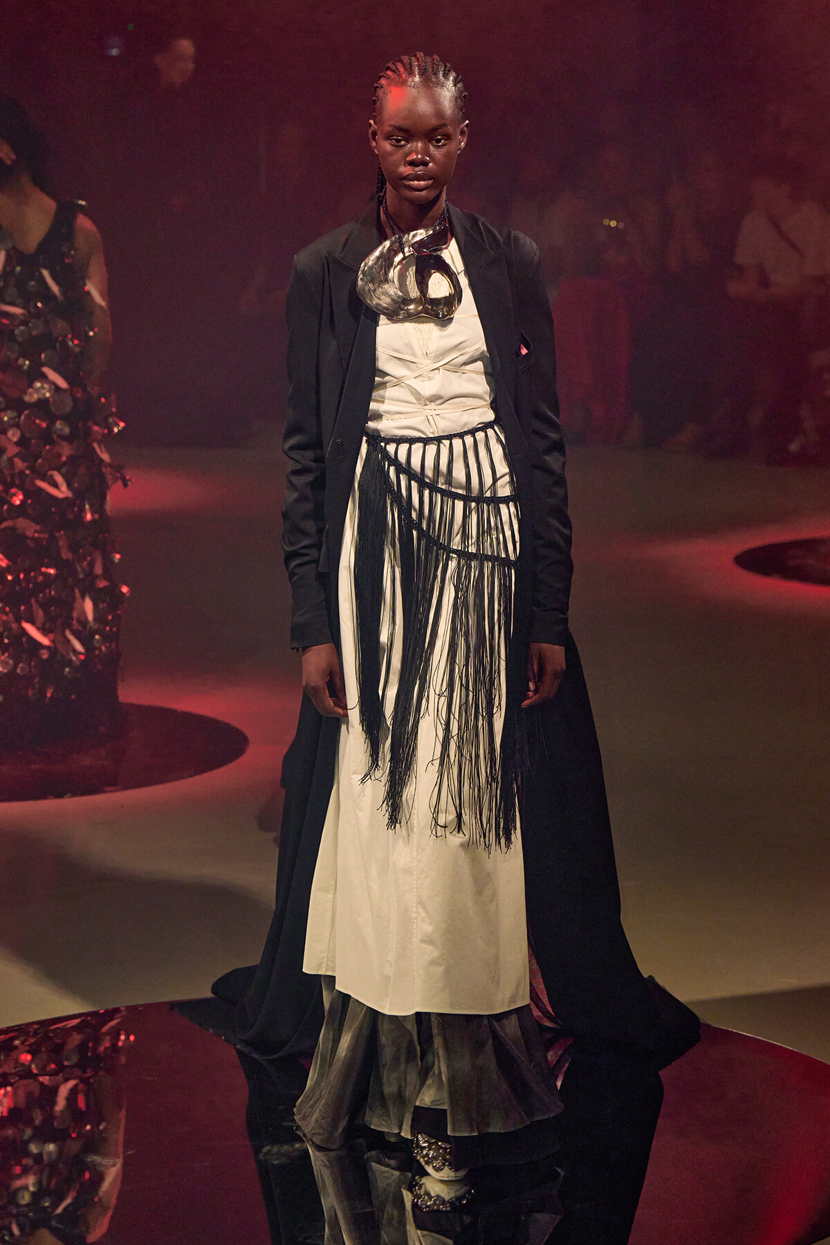 Runway model in a white dress with black fringe, wearing a long black coat and a bold metallic necklace under dramatic lighting.