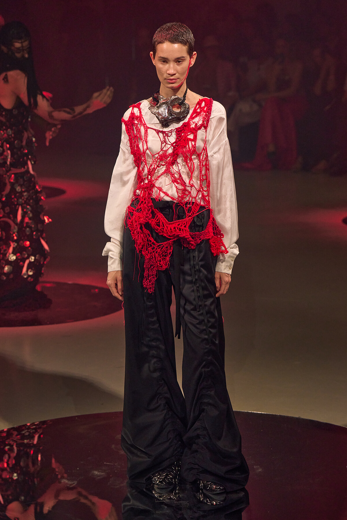 Model on a runway wearing a white blouse covered in red crocheted netting over black wide-leg pants, with a dark statement necklace at the neck.
