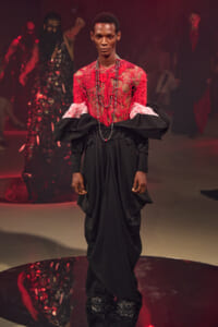 Male model on a runway wearing a red crochet top with a black draped skirt and layered bead necklaces.