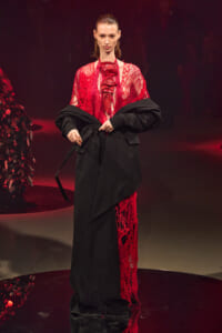 Model on a runway wearing a red lace top and a black oversized coat.