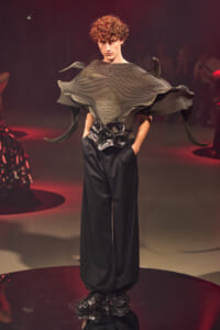 Model on a fashion runway wearing a sculptural metallic gray top with wing-like ruffles and wide black trousers.
