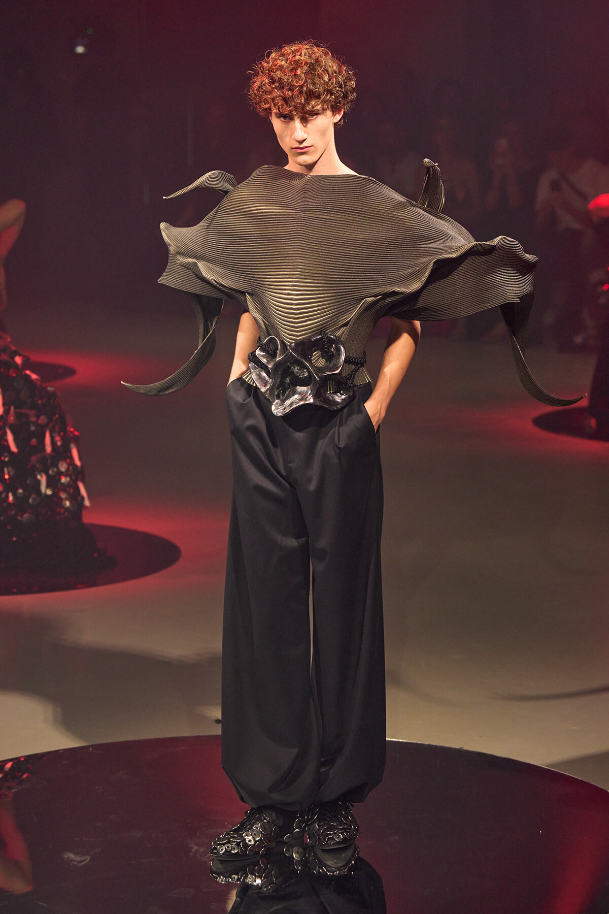 Model on a fashion runway wearing a sculptural metallic gray top with wing-like ruffles and wide black trousers.