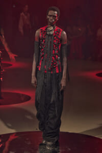 Male model walks the runway in a dramatic red-and-black embellished outfit with long black fringe, standing on stage.