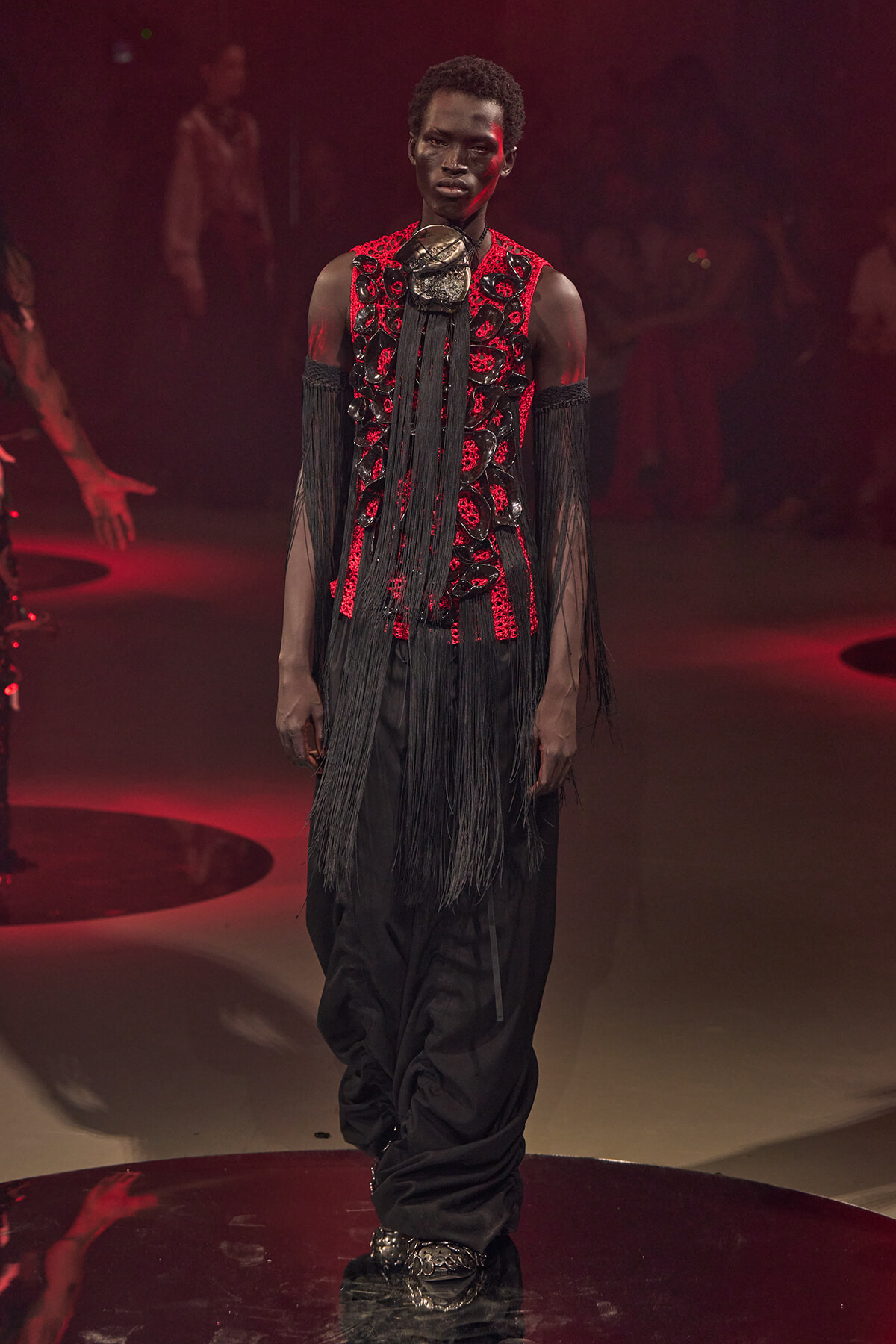 Male model walks the runway in a dramatic red-and-black embellished outfit with long black fringe, standing on stage.