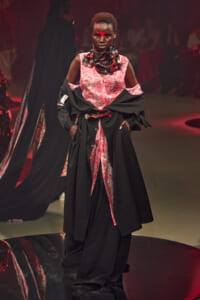 Model on a runway wearing a pink patterned dress with a black overcoat, hands in pockets, red face paint across the nose, dramatic scarf around neck