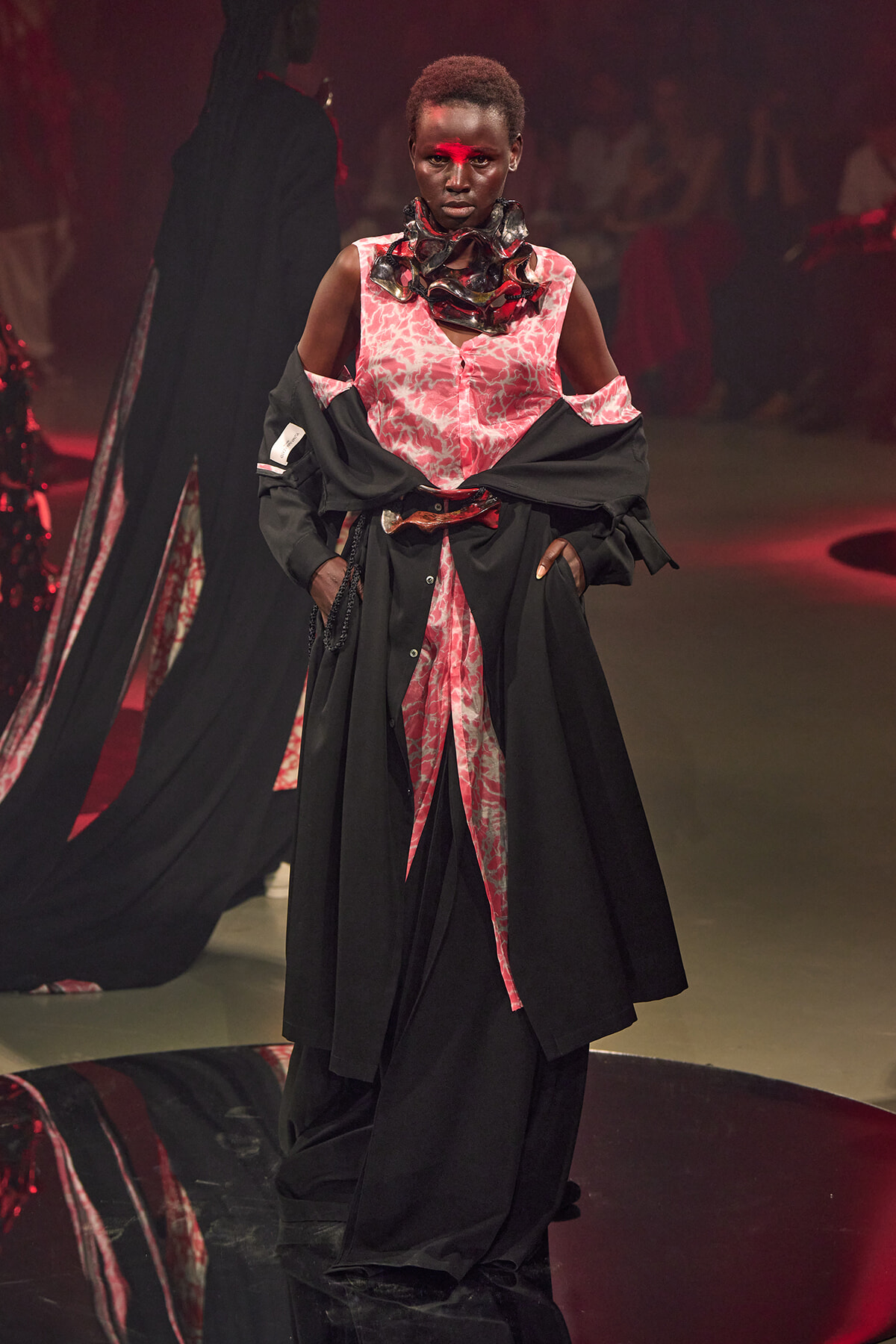 Model on a runway wearing a pink patterned dress with a black overcoat, hands in pockets, red face paint across the nose, dramatic scarf around neck