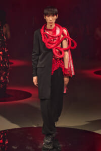 Model on a runway wearing a black suit with a bold red rope sculpture draped over the shoulder and chest.