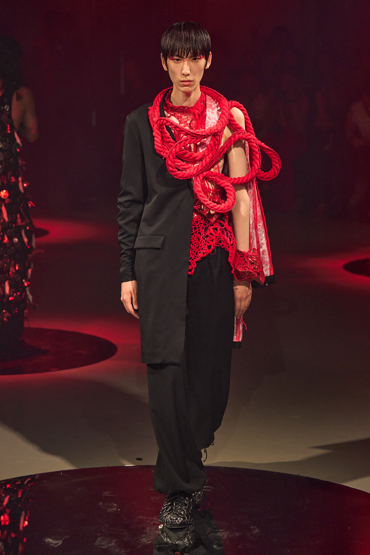 Model on a runway wearing a black suit with a bold red rope sculpture draped over the shoulder and chest.