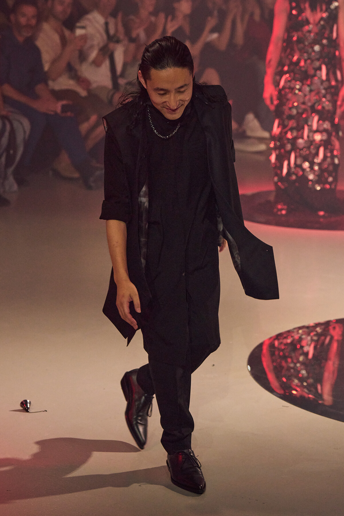 Model walking a fashion runway in an all-black outfit under red lighting, audience in the background. They bow slightly with one foot forward and a long coat draped over shoulders.