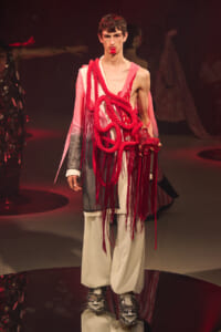 Male model on a runway wearing a white suit with a red rope harness and dangling fringe details; bold red accents dominate the look.