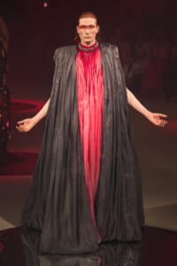 Model on a runway wearing a voluminous black cape over a bright pink sheer gown with bold red eye makeup and a beaded collar.