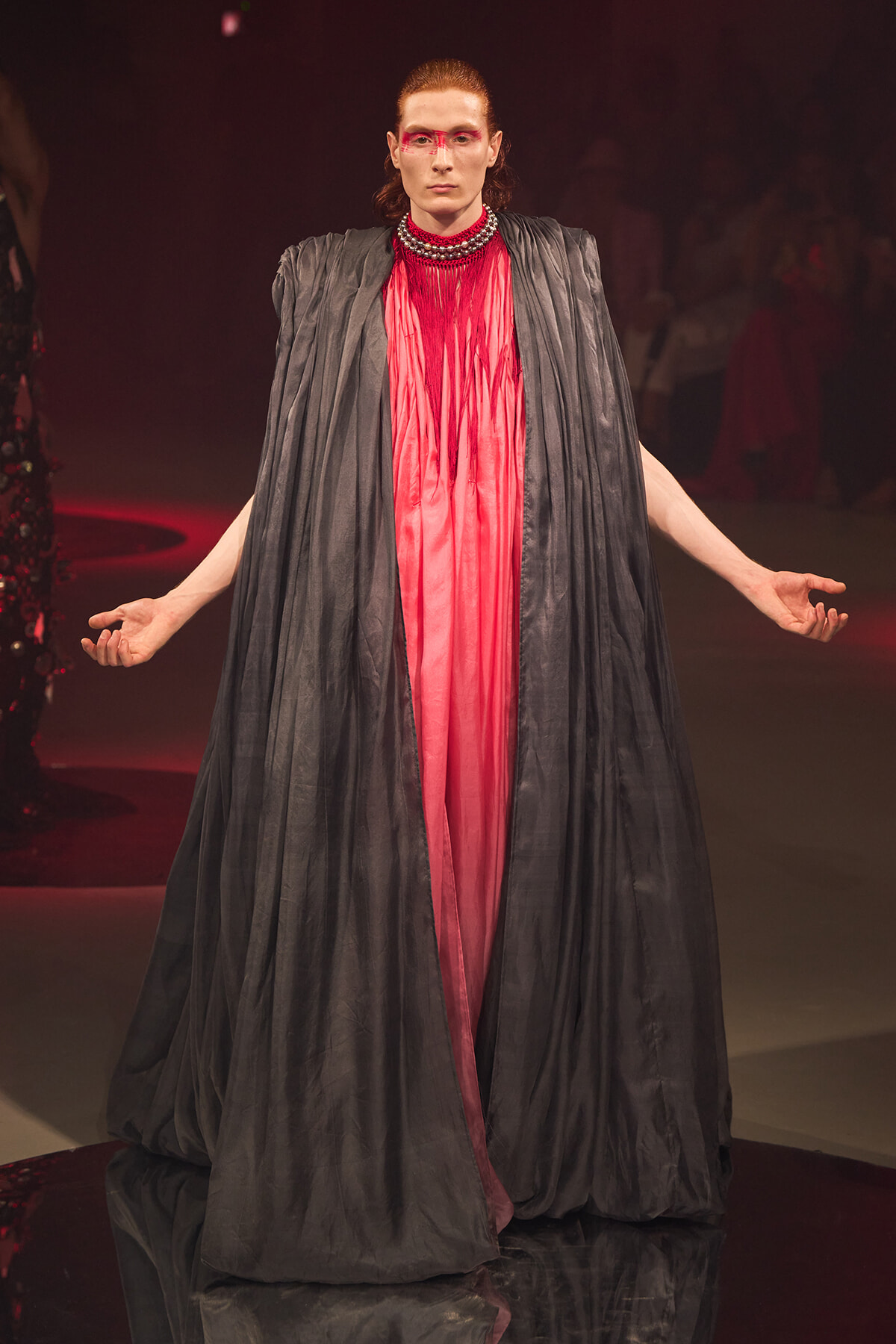 Model on a runway wearing a voluminous black cape over a bright pink sheer gown with bold red eye makeup and a beaded collar.