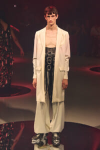 Male model on a fashion runway wearing a cream oversized blazer over a black beaded fringe top and wide white trousers with embellished platform shoes.