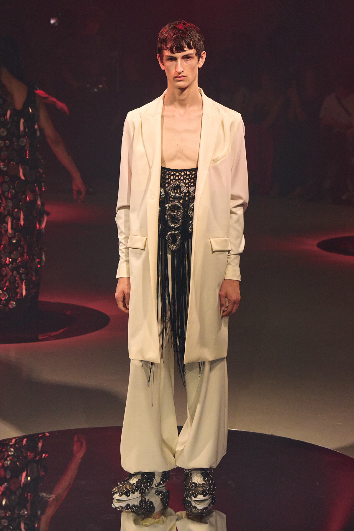 Male model on a fashion runway wearing a cream oversized blazer over a black beaded fringe top and wide white trousers with embellished platform shoes.