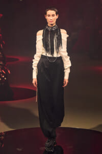 Model on a dim runway wearing an off-shoulder white blouse with a black fringe bib and long necklace over voluminous black pants, black boots visible at the bottom.