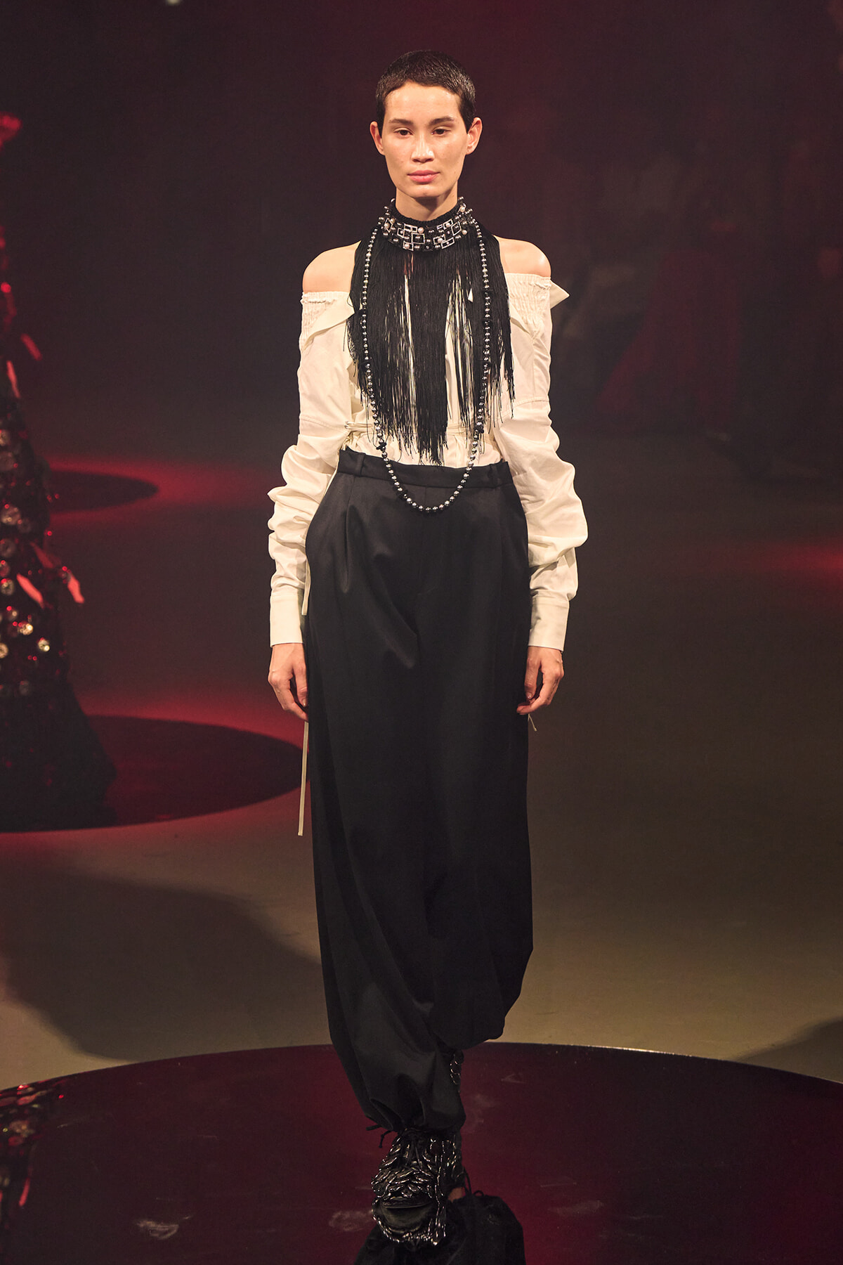 Model on a dim runway wearing an off-shoulder white blouse with a black fringe bib and long necklace over voluminous black pants, black boots visible at the bottom.