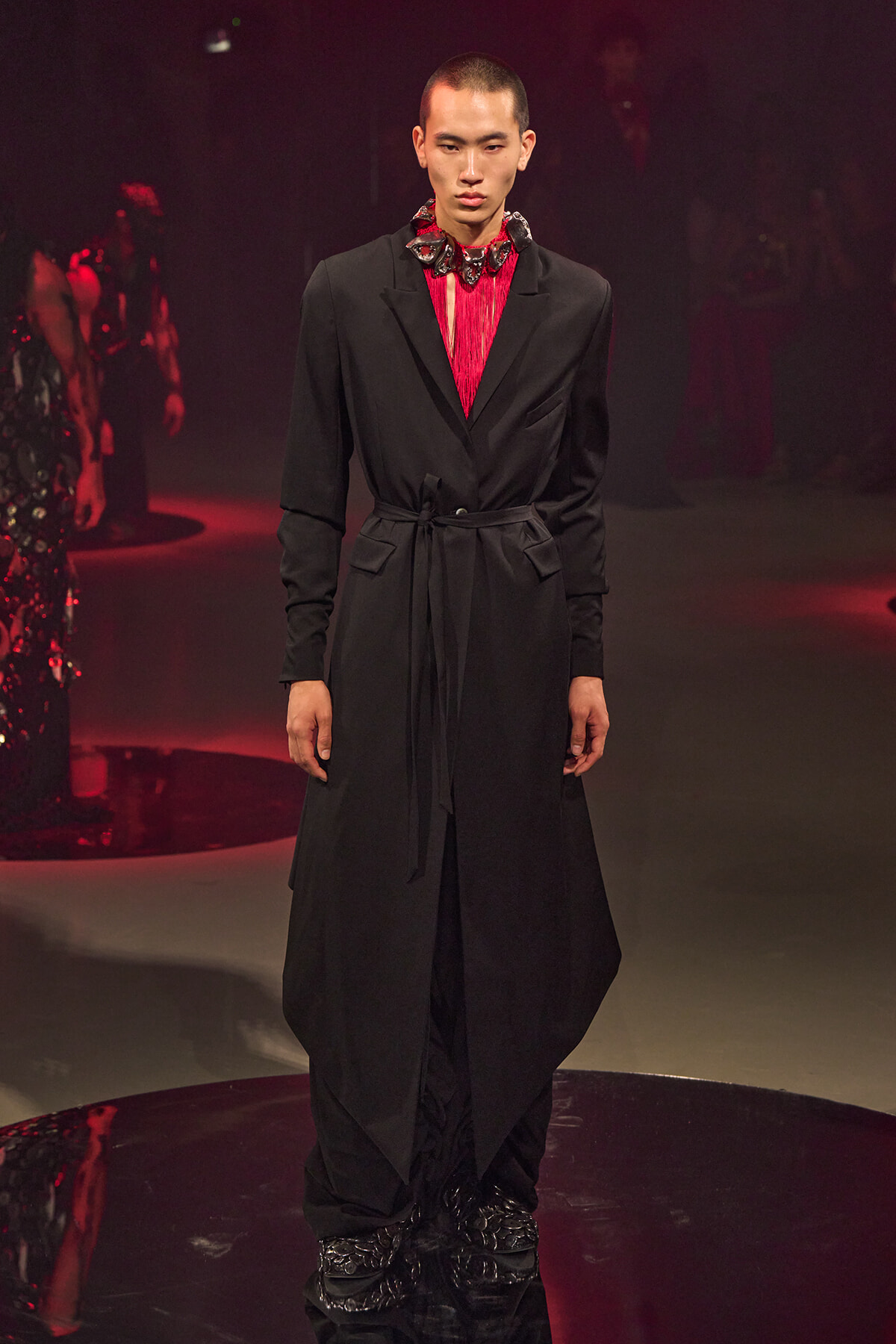 Male model walking the runway in a long black belted coat over a red top with a decorative neckpiece, under dramatic red-lit lighting.