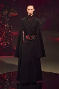 Model on a runway wearing a long black coat with a red-black floral belt and ruffled neck detail.