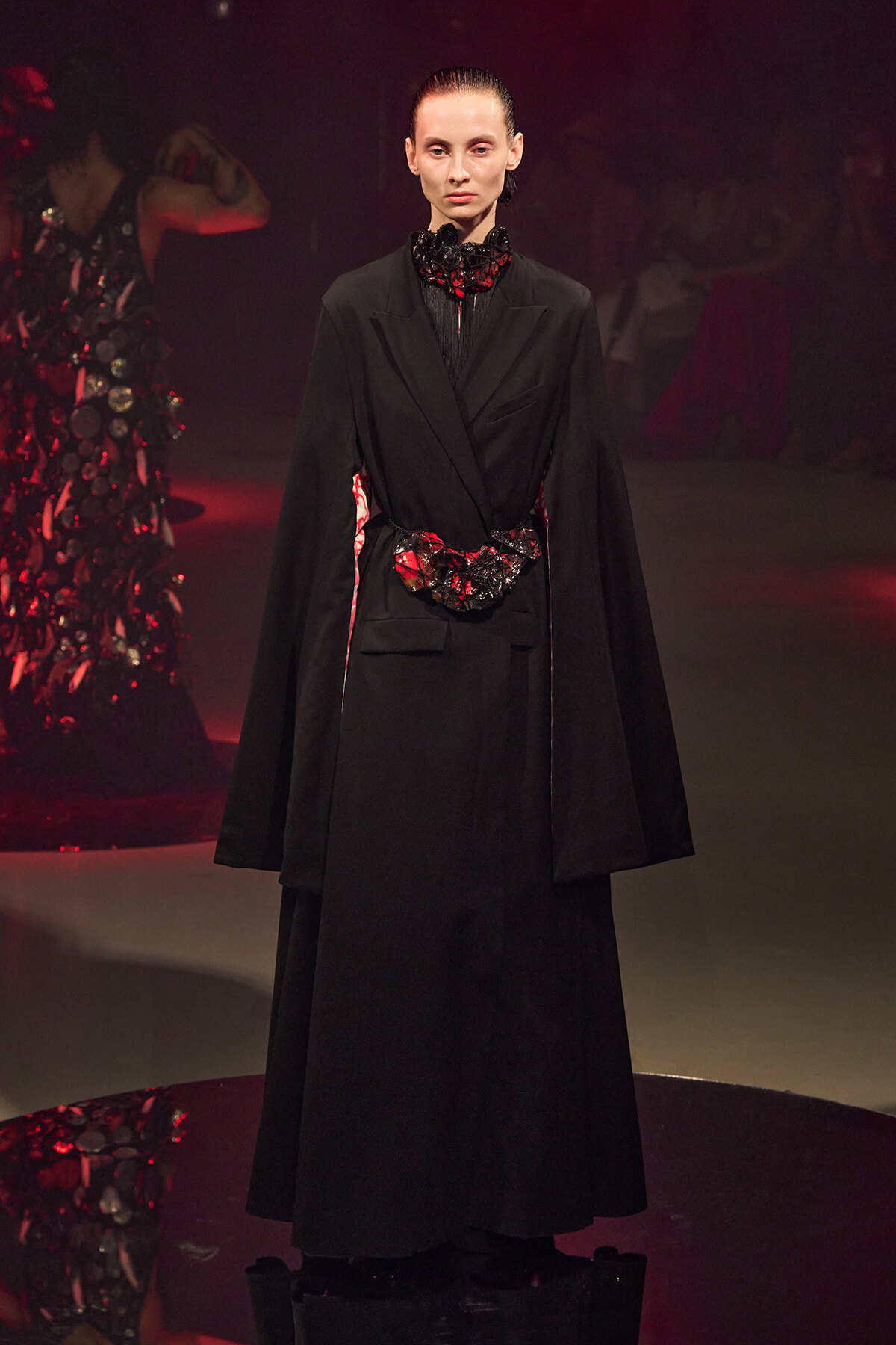 Model on a runway wearing a long black coat with a red-black floral belt and ruffled neck detail.