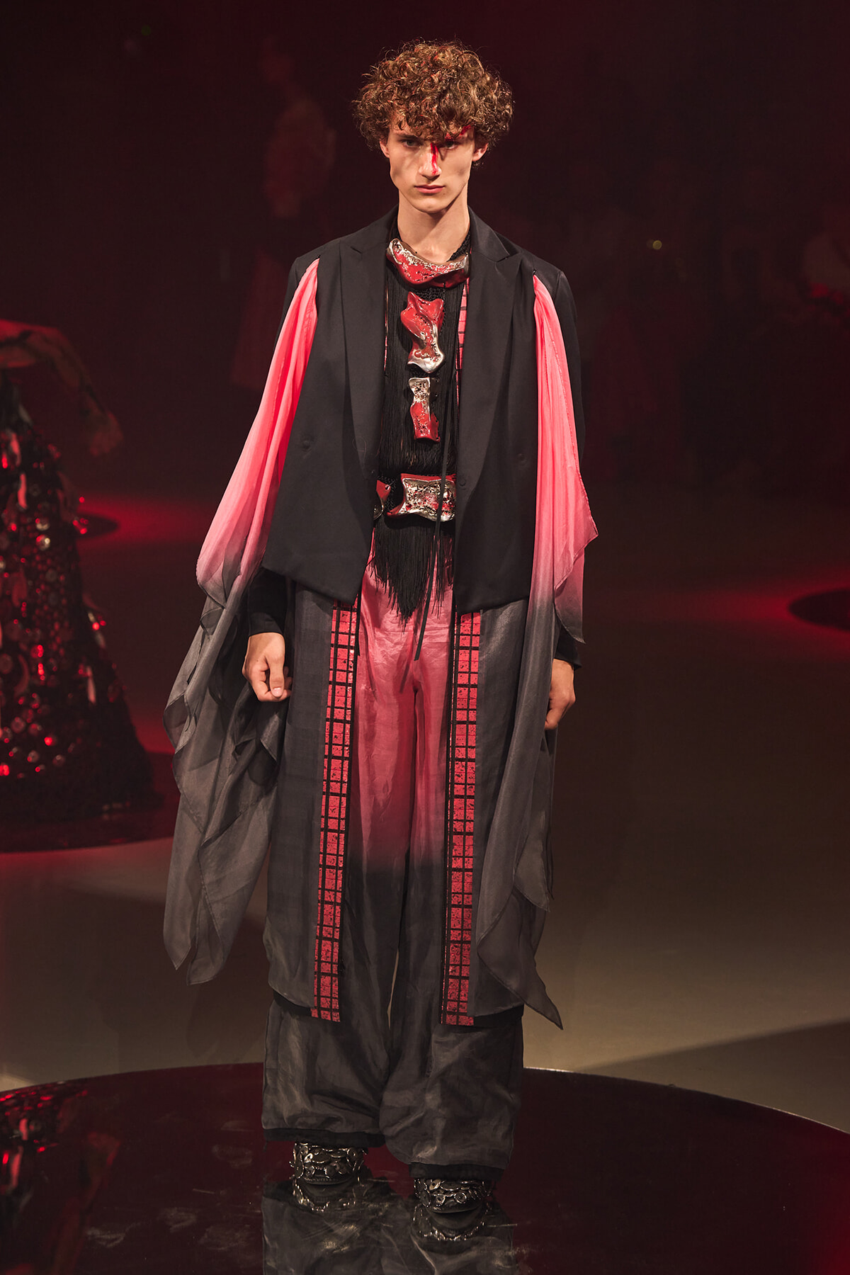 Male model walks a fashion show wearing a black sleeveless coat over a pink-to-red gradient robe with bold metallic neck pieces.