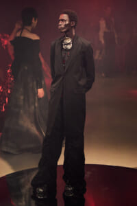 Male model in a long black coat with a large metallic necklace on a red-lit runway.