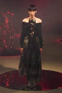 Model on a runway wearing an off-the-shoulder black top and long, dark skirt with a sculptural metallic neckpiece under red stage lighting.