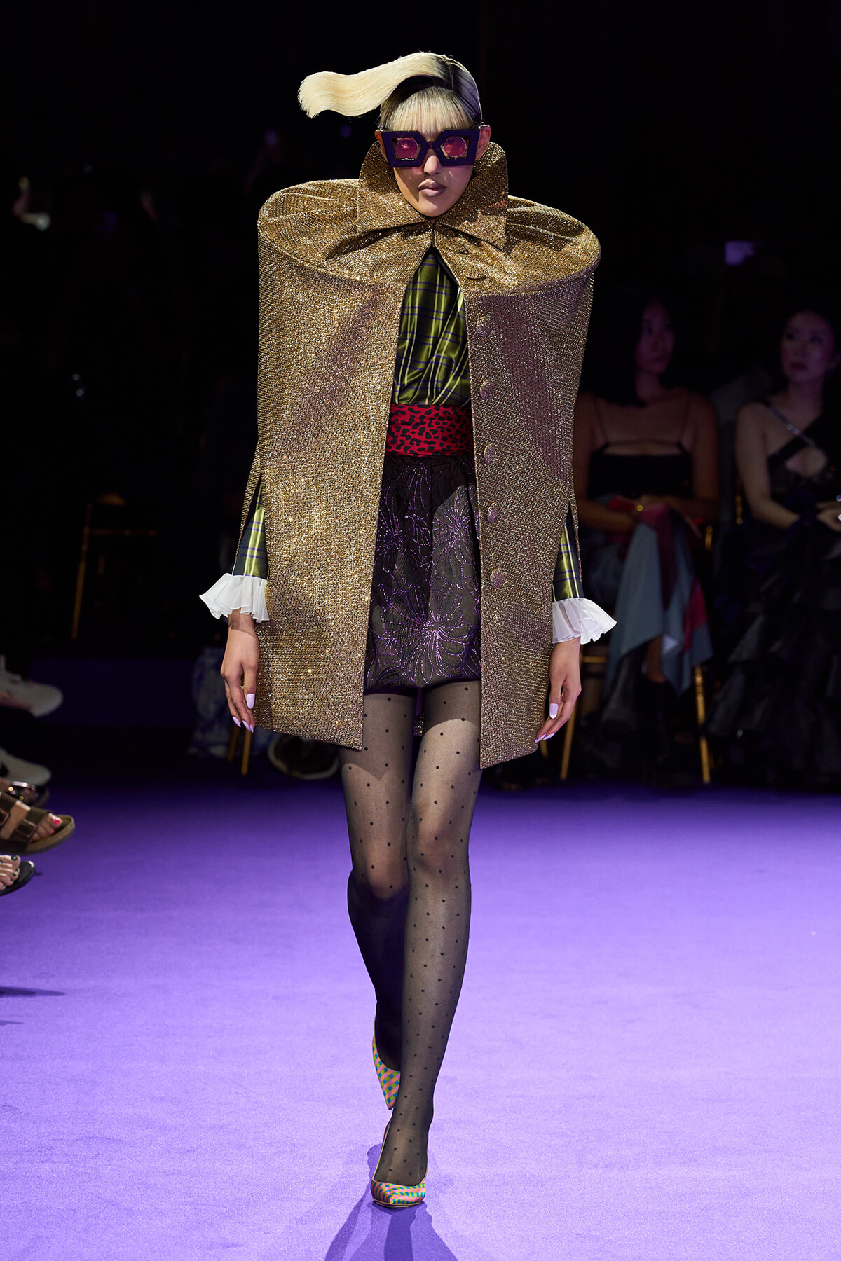 Runway model in a shimmering gold cape over a plaid green-and-black outfit, dotted tights, and colorful heels, wearing oversized purple sunglasses.