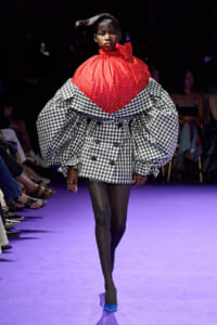 Runway model wears a dramatic red polka-dot top over a voluminous black-and-white gingham coat with puff sleeves during a fashion show.