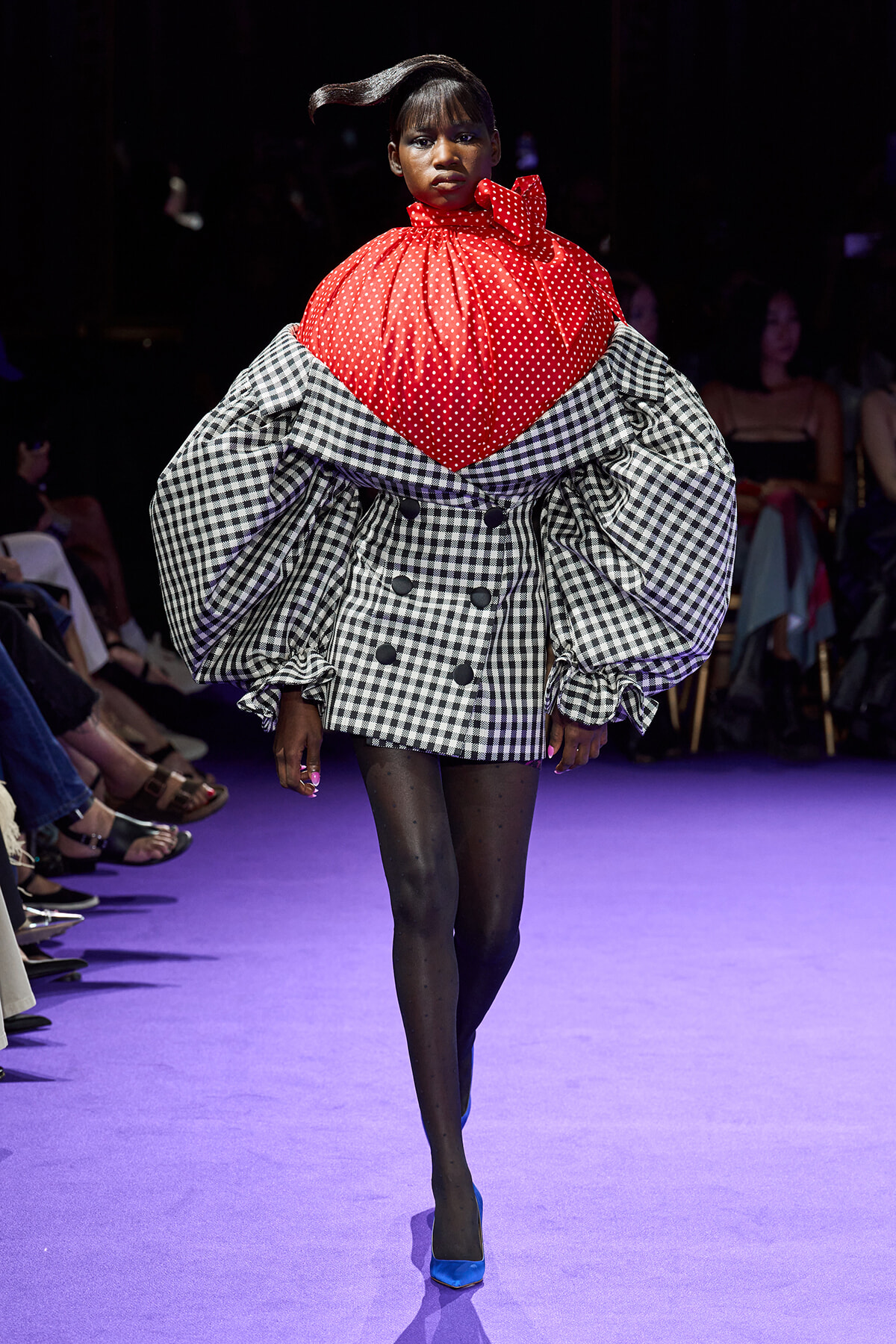 Runway model wears a dramatic red polka-dot top over a voluminous black-and-white gingham coat with puff sleeves during a fashion show.