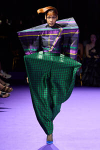Model on a purple-lit runway wearing a sculptural purple-striped top and oversized green houndstooth pants.
